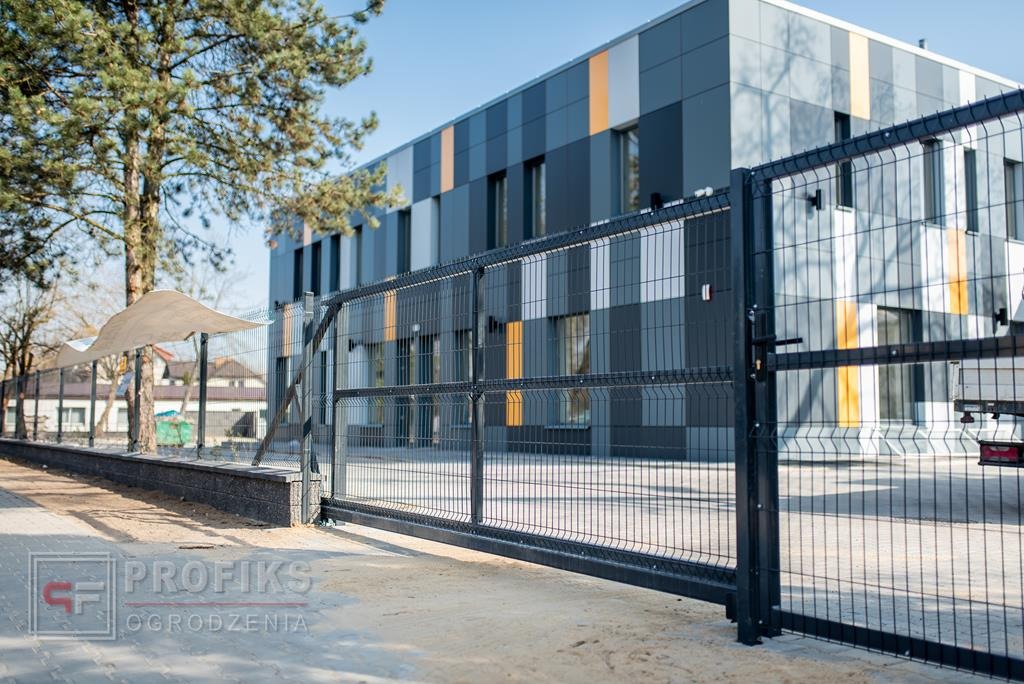 Przesuwna brama z paneli z widocznym mechanizmem jezdnym na tle nowoczesnego budynku z panelami w odcieniach szarości i pomarańczy.