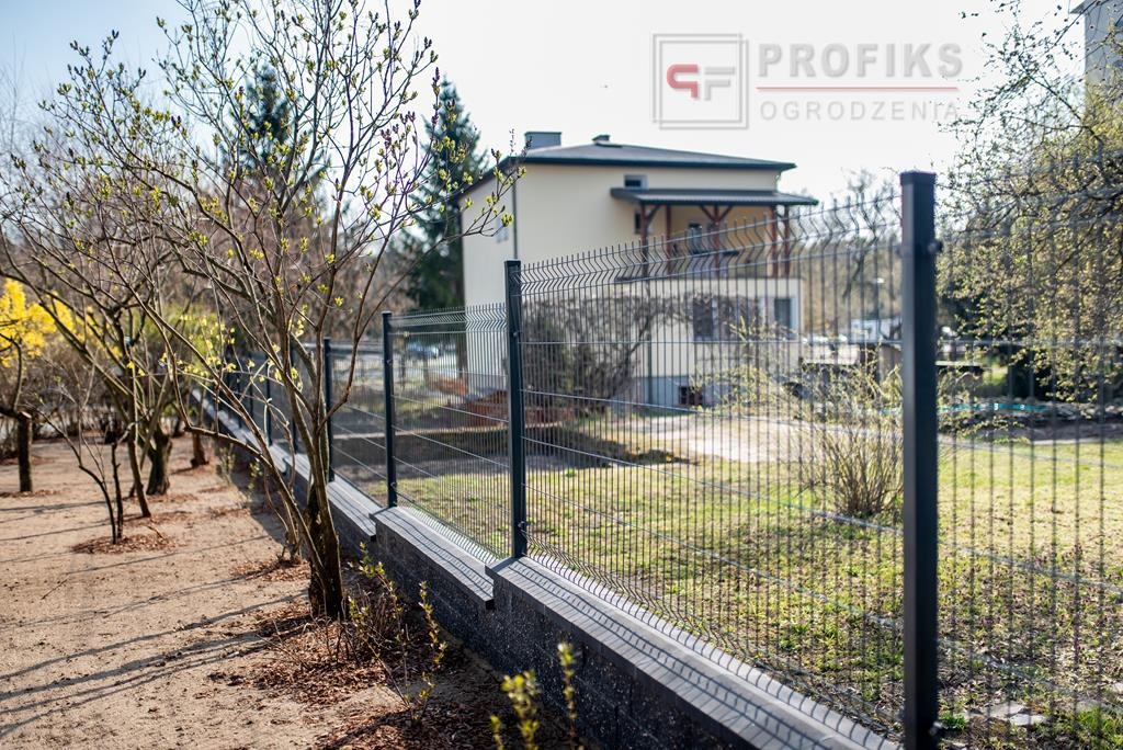 Nowoczesne, grafitowe ogrodzenie panelowe na betonowej podmurówce, widok na dom w tle, drzewa z pąkami na wiosnę.