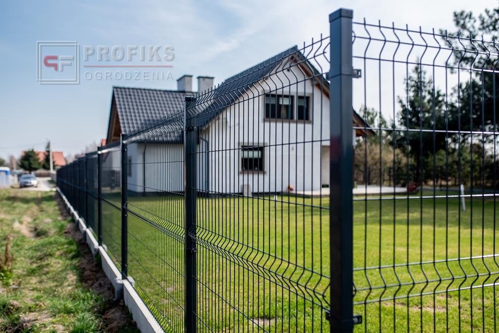 Nowoczesne ogrodzenie panelowe w kolorze grafitowym, z widocznym betonowym podmurówką i domem w tle, w słoneczny dzień.