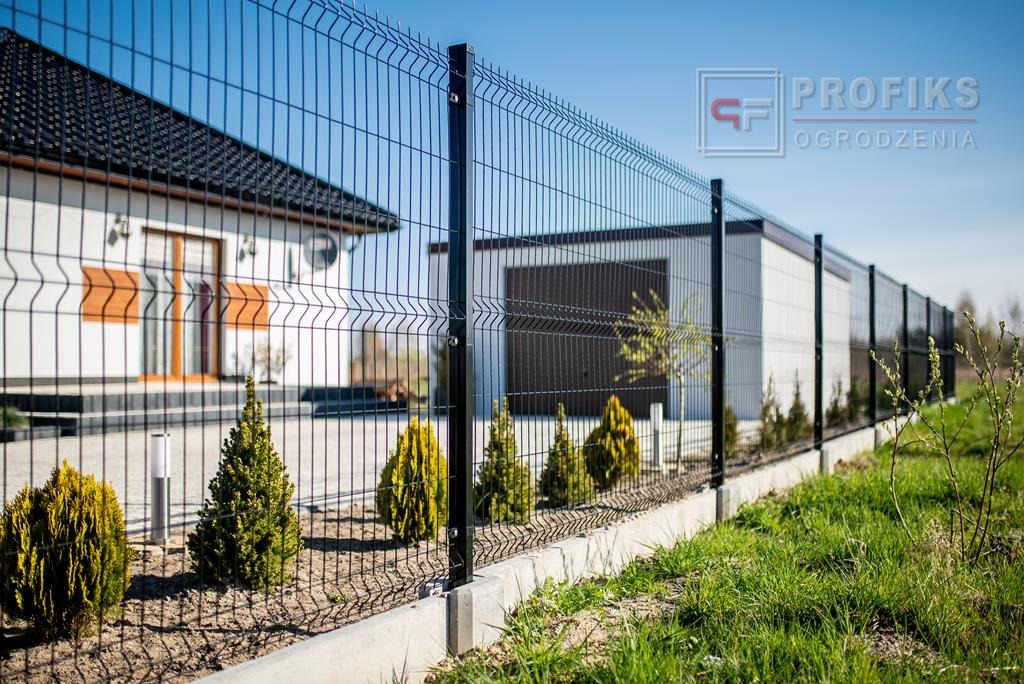 Długi, czarny system ogrodzeniowy z siatki panelowej, zamontowany na betonowej podmurówce, oddzielający posesję z domem i garażem, z widocznymi młodymi krzewami ozdobnymi.