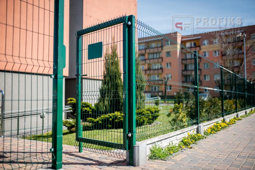 Otwarta, zielona furtka z siatki panelowej, z widocznym zamkiem i słupkami, prowadząca na posesję z niską roślinnością i budynkiem mieszkalnym w tle.