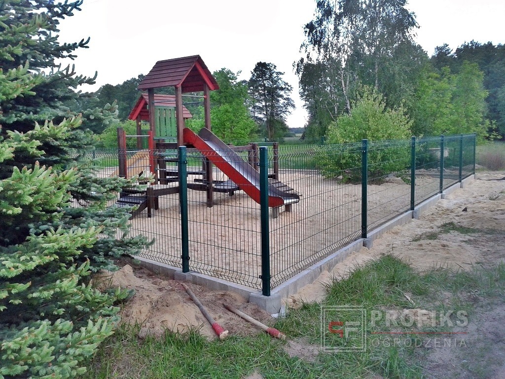 Zielone, metalowe ogrodzenie wokół placu zabaw z piaskiem, widoczne narzędzia i logo firmy w prawym dolnym rogu.