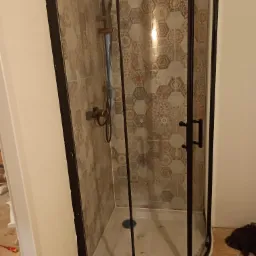 Narożna kabina prysznicowa z czarną ramą i dekoracyjnymi, heksagonalnymi płytkami wewnątrz, zamontowana w trakcie remontu łazienki, widoczne niedokończone podłogi.