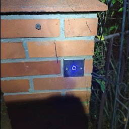 Bartosz K. Volt System - Przycisk domofonu z niebieskim podświetleniem zamontowany na ceglanym słupku bramy, widoczne elementy ogrodzenia i roślinność w tle.