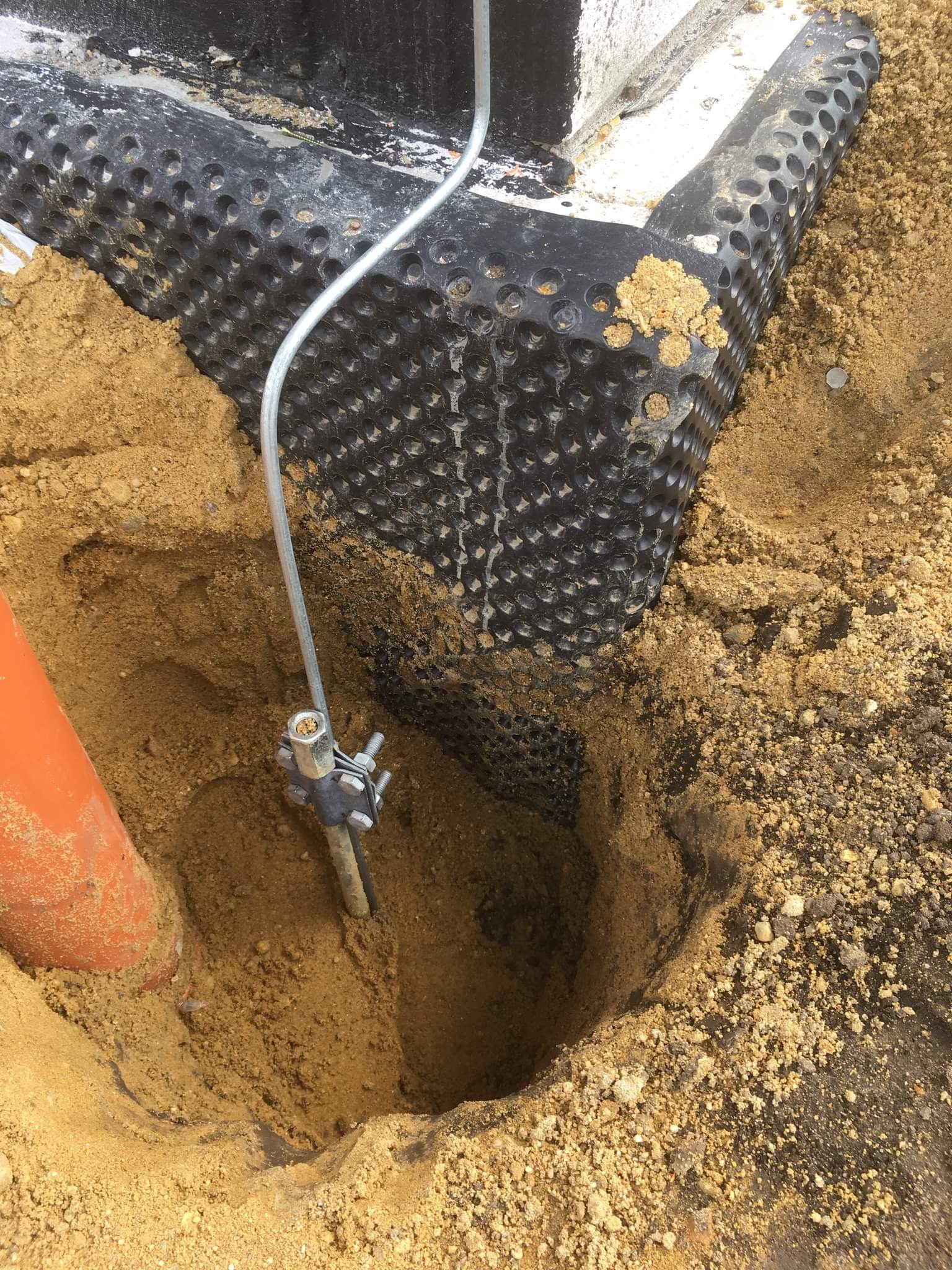 Uziemienie pionowe w wykopie, widoczna czarna folia kubełkowa przy fundamencie, pomarańczowa rura kanalizacyjna w piasku.