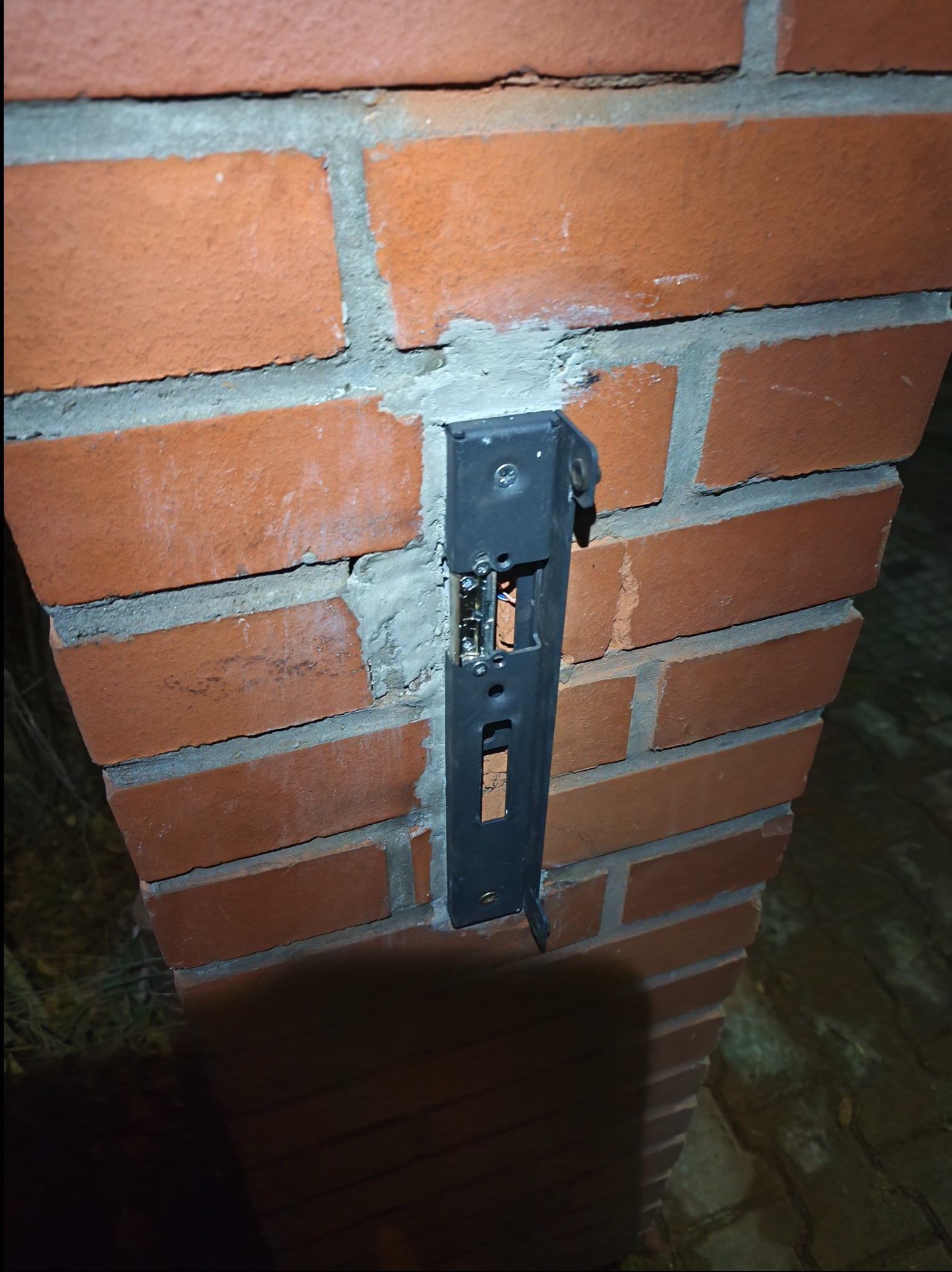 Ciemne ujęcie montażu wideodomofonu na ceglanym słupku. Widoczne okablowanie wewnątrz metalowej obudowy. Detal montażu na zewnątrz budynku.