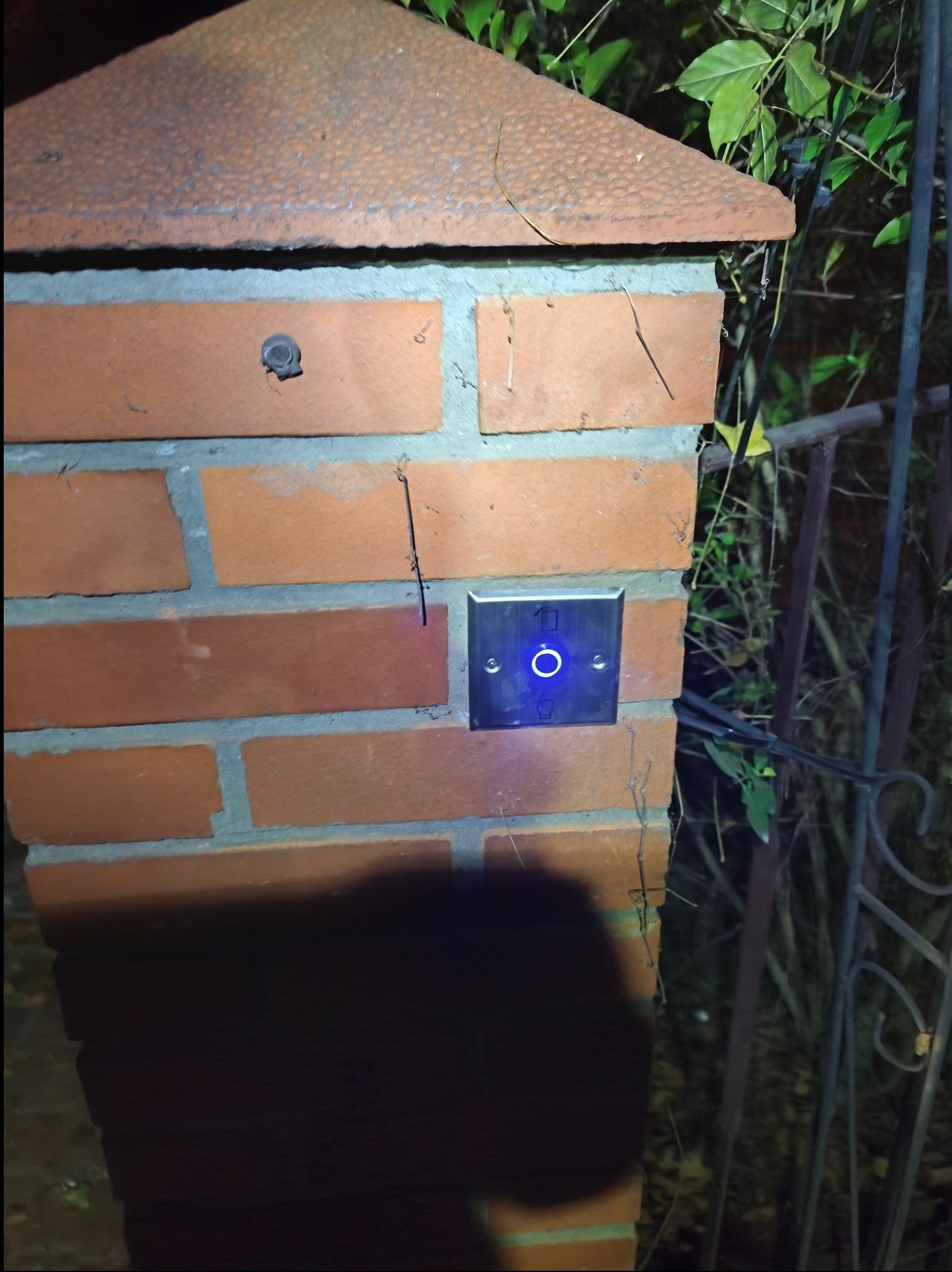 Przycisk domofonu z niebieskim podświetleniem zamontowany na ceglanym słupku bramy, widoczne elementy ogrodzenia i roślinność w tle.