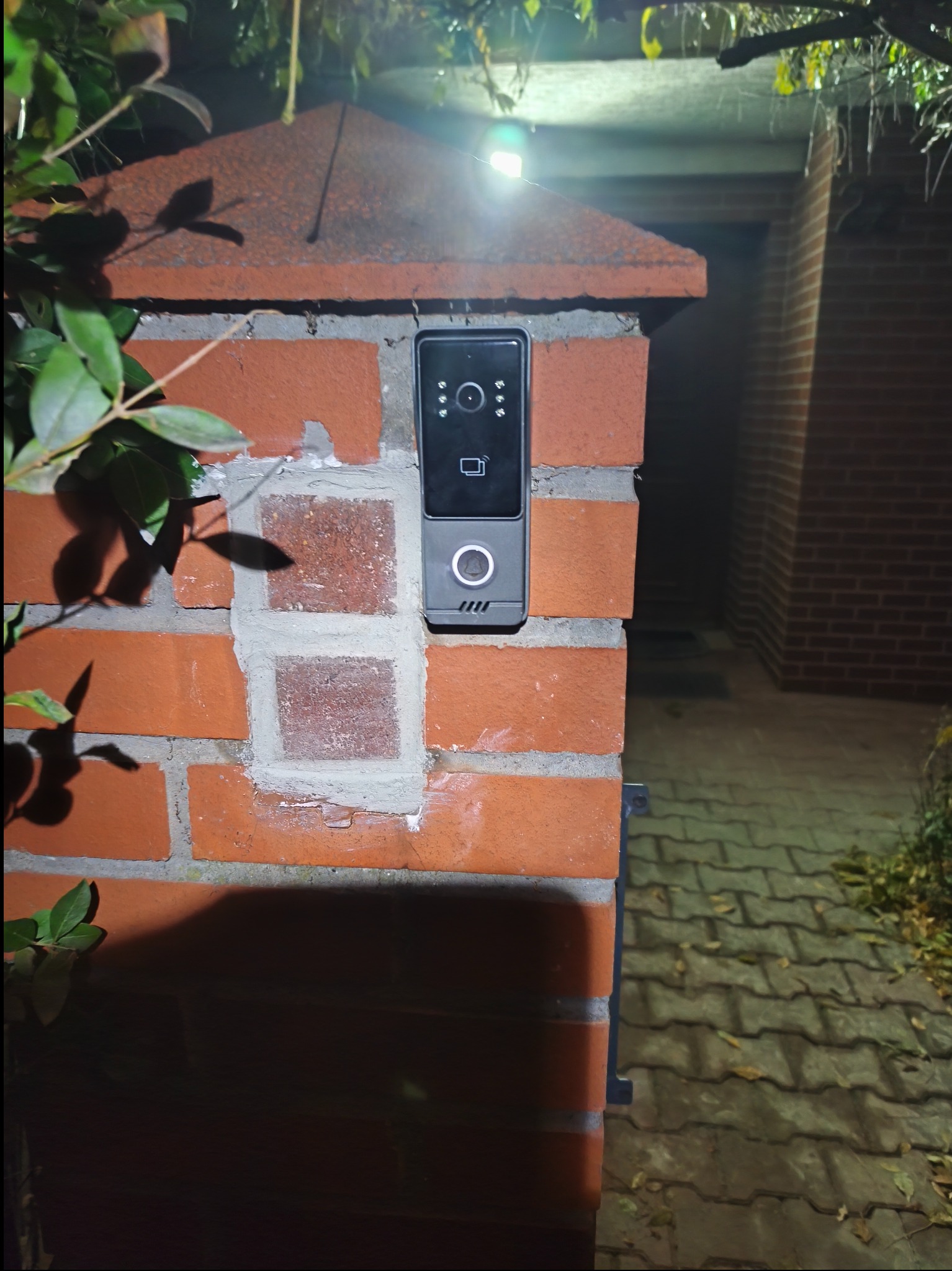 Nowoczesny wideodomofon zamontowany na ceglanym słupku bramy wjazdowej, oświetlony górnym światłem. Widoczne detale montażu i otoczenie bramy.