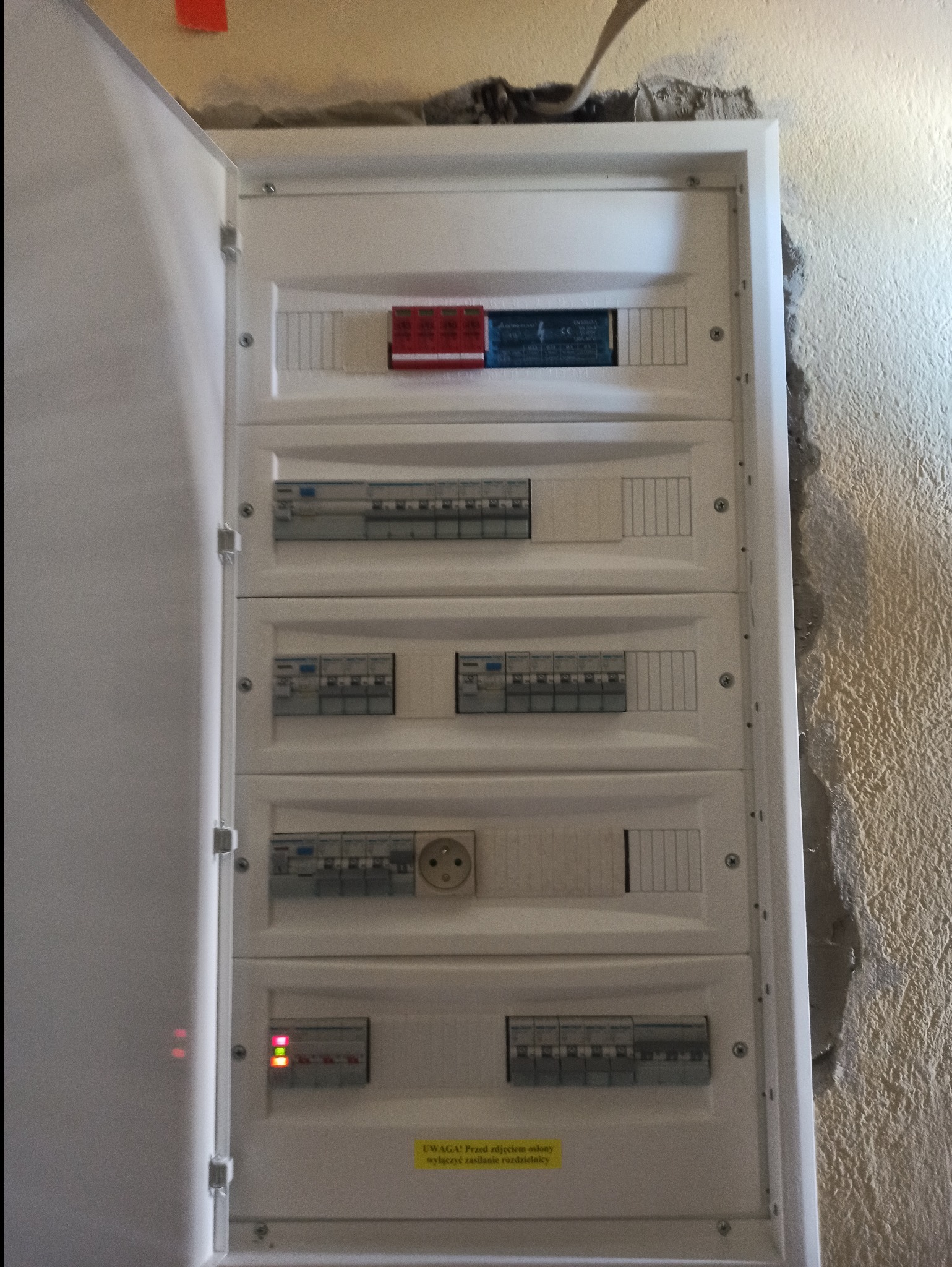 Otwarta, biała skrzynka rozdzielcza z bezpiecznikami, gniazdkiem i żółtą naklejką ostrzegawczą na tle ściany o nierównej fakturze. Widoczne elementy instalacji elektrycznej.