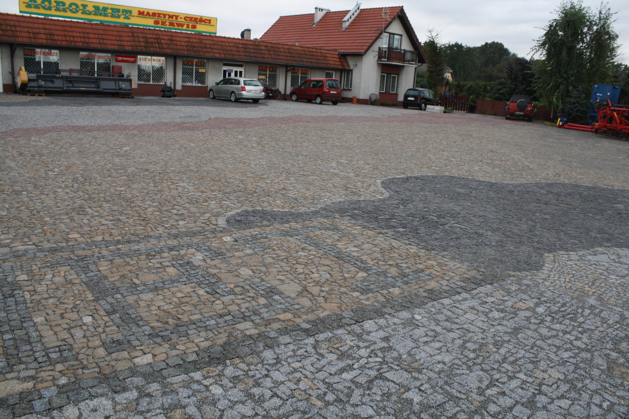 Rozległy plac wyłożony kostką granitową w różnych odcieniach szarości i beżu, z widocznym wzorem i fragmentem ciemniejszego obszaru, budynki w tle.