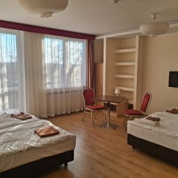 PP DP DMYTRO PYROHOV - Jasny pokój hotelowy z dwoma łóżkami, stolikiem i krzesłami. Duże okna z białymi zasłonami wpuszczają naturalne światło. Widoczne półki w ścianie i telewizor.