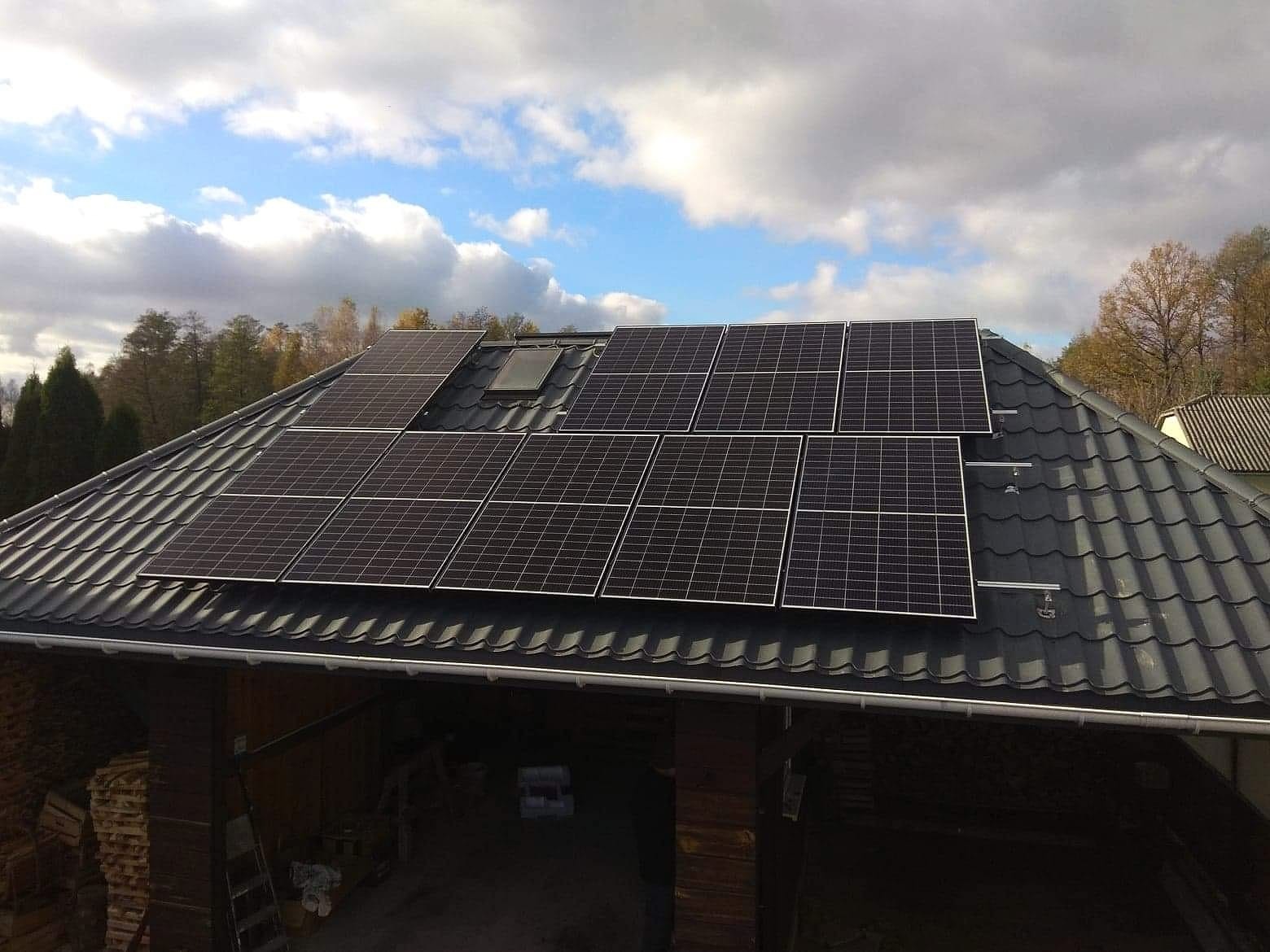 Panele słoneczne na dachu domu z ciemną dachówką, widoczne mocowania paneli, w tle błękitne niebo z chmurami i drzewa.