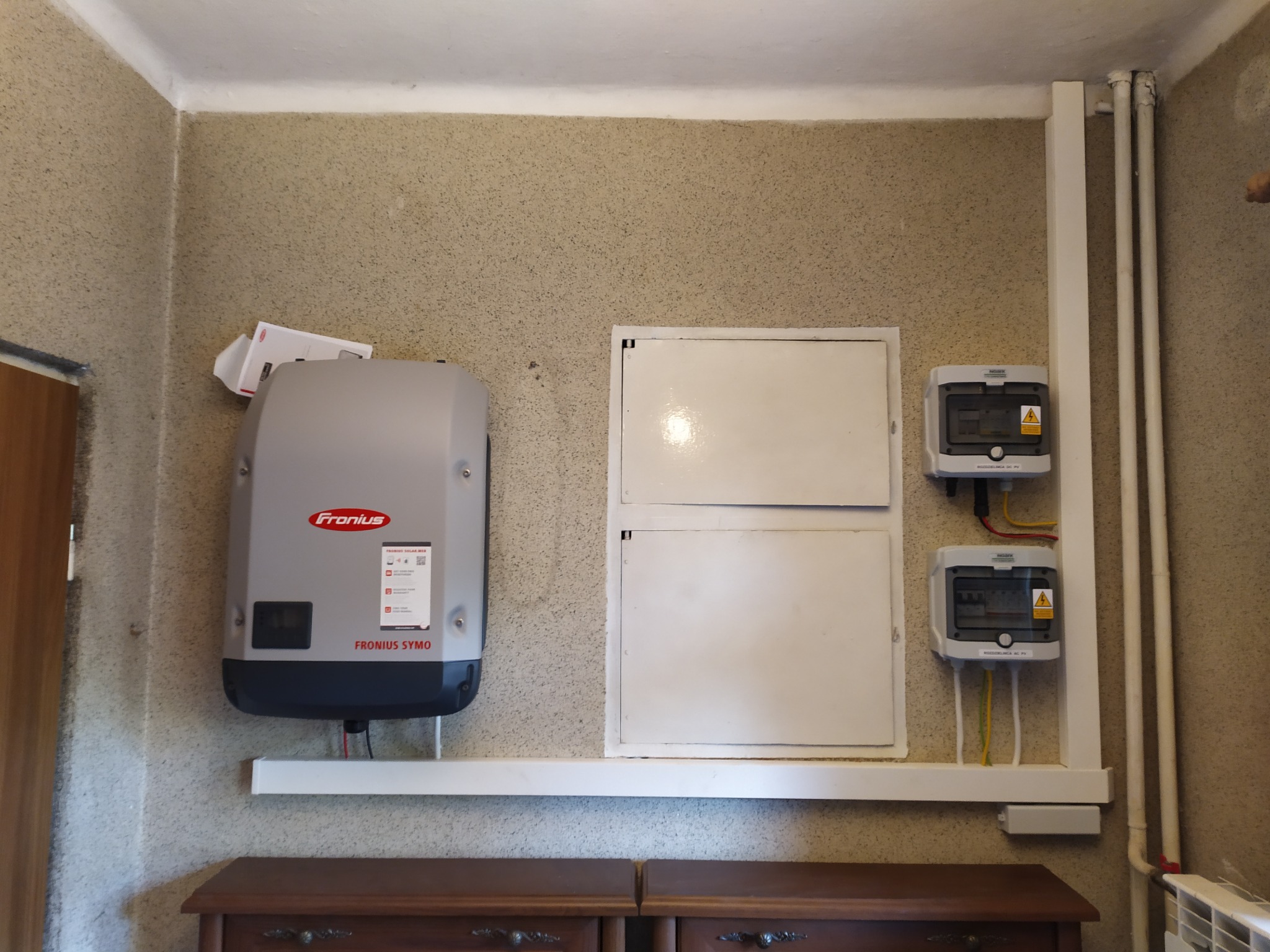 Zamontowany inwerter Fronius Symo na ścianie o fakturze baranka, skrzynki elektryczne i rury grzewcze w tle, nad starą komodą.