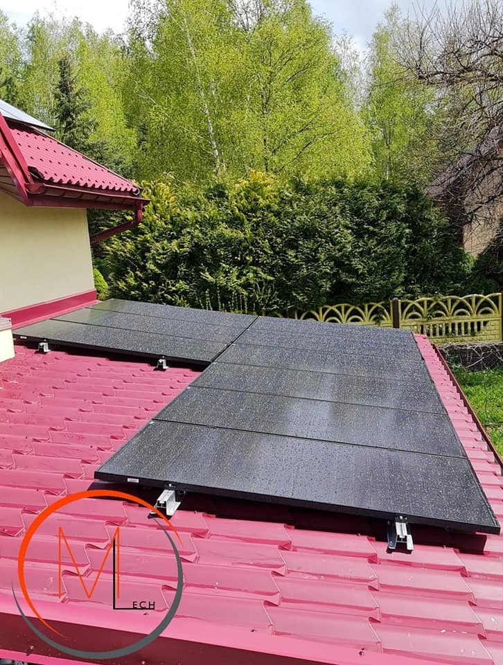 Panele słoneczne na czerwonym dachu pokrytym kroplami deszczu, widoczne elementy montażowe i logo firmy w rogu zdjęcia.