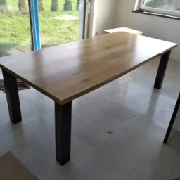 Stół z blatem z jasnego drewna i czarnymi, kwadratowymi nogami, ustawiony w jasnym pomieszczeniu z dużym oknem z widokiem na zieleń. Widoczny fragment podłogi z płytek i elementy wykończenia wnętrza.