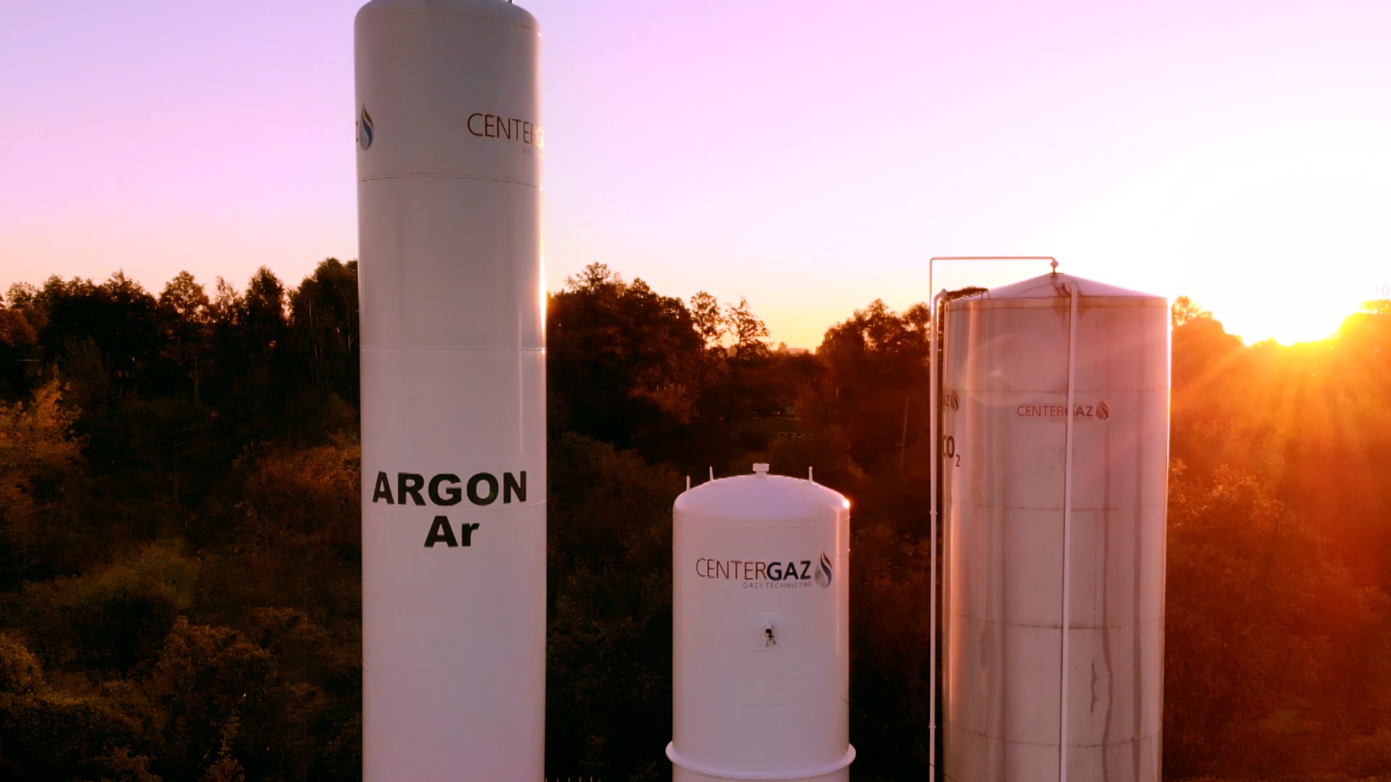 Trzy białe zbiorniki gazowe różnych rozmiarów z logo firmy na tle zachodzącego słońca i drzew