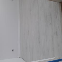 Bez nazwy - Świeżo pomalowana, biała ściana w rogu pomieszczenia z widocznym gniazdkiem elektrycznym oraz jasna podłoga imitująca drewno, częściowo zabezpieczona taśmą malarską.