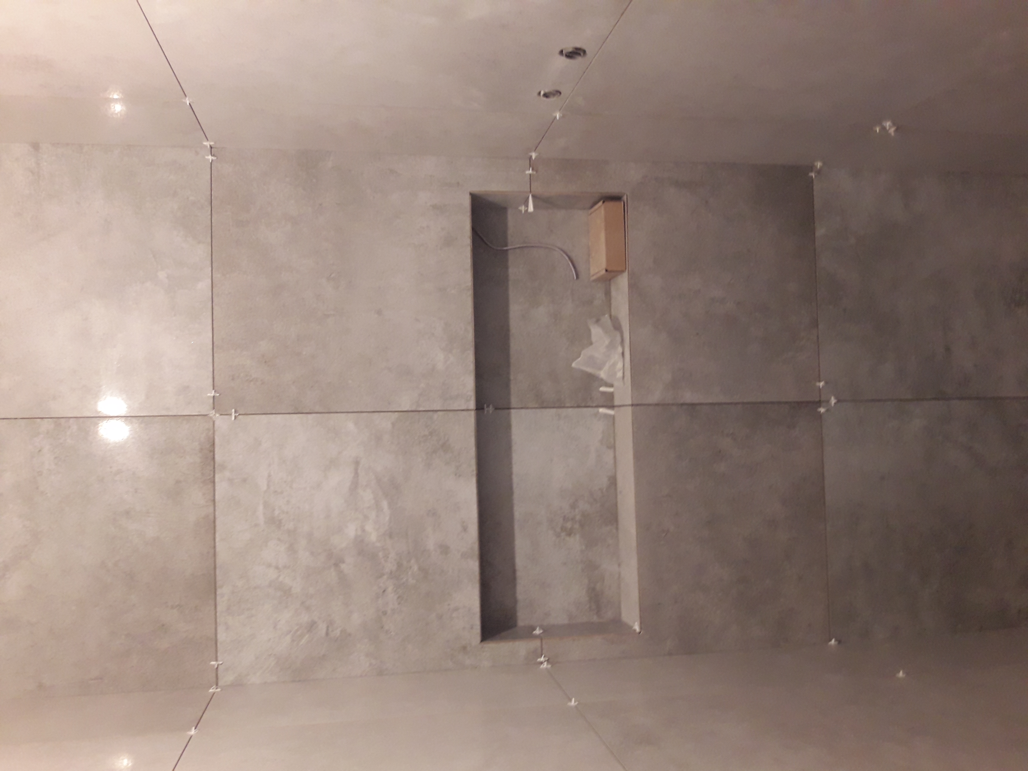 Ściana i sufit w trakcie wykańczania z szarymi płytkami, widoczne krzyżyki dystansowe i wnęka z wystającym kablem.