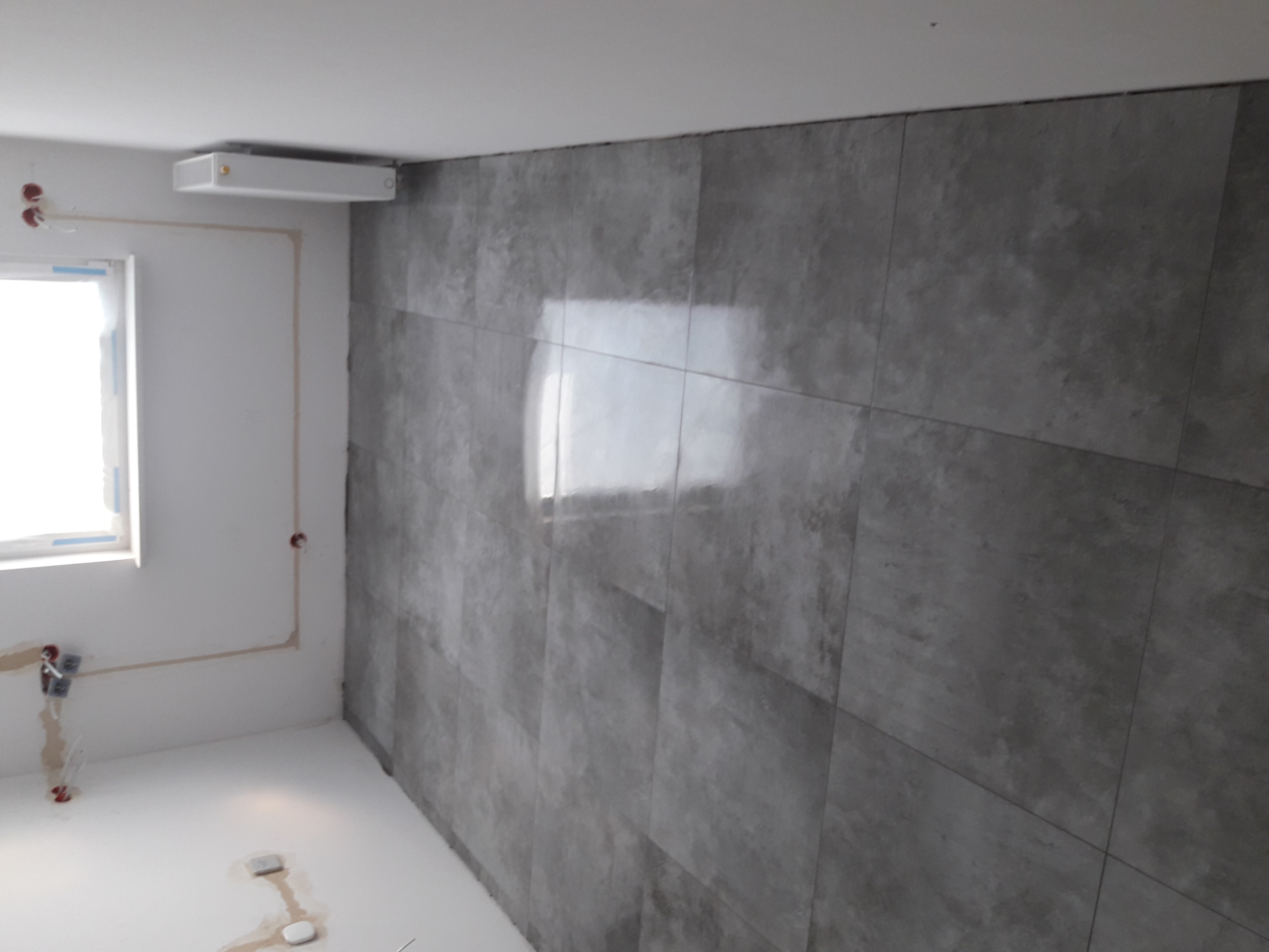 Świeżo wykończone wnętrze z szarymi płytkami imitującymi beton na ścianie, widoczne puszki elektryczne i okno.