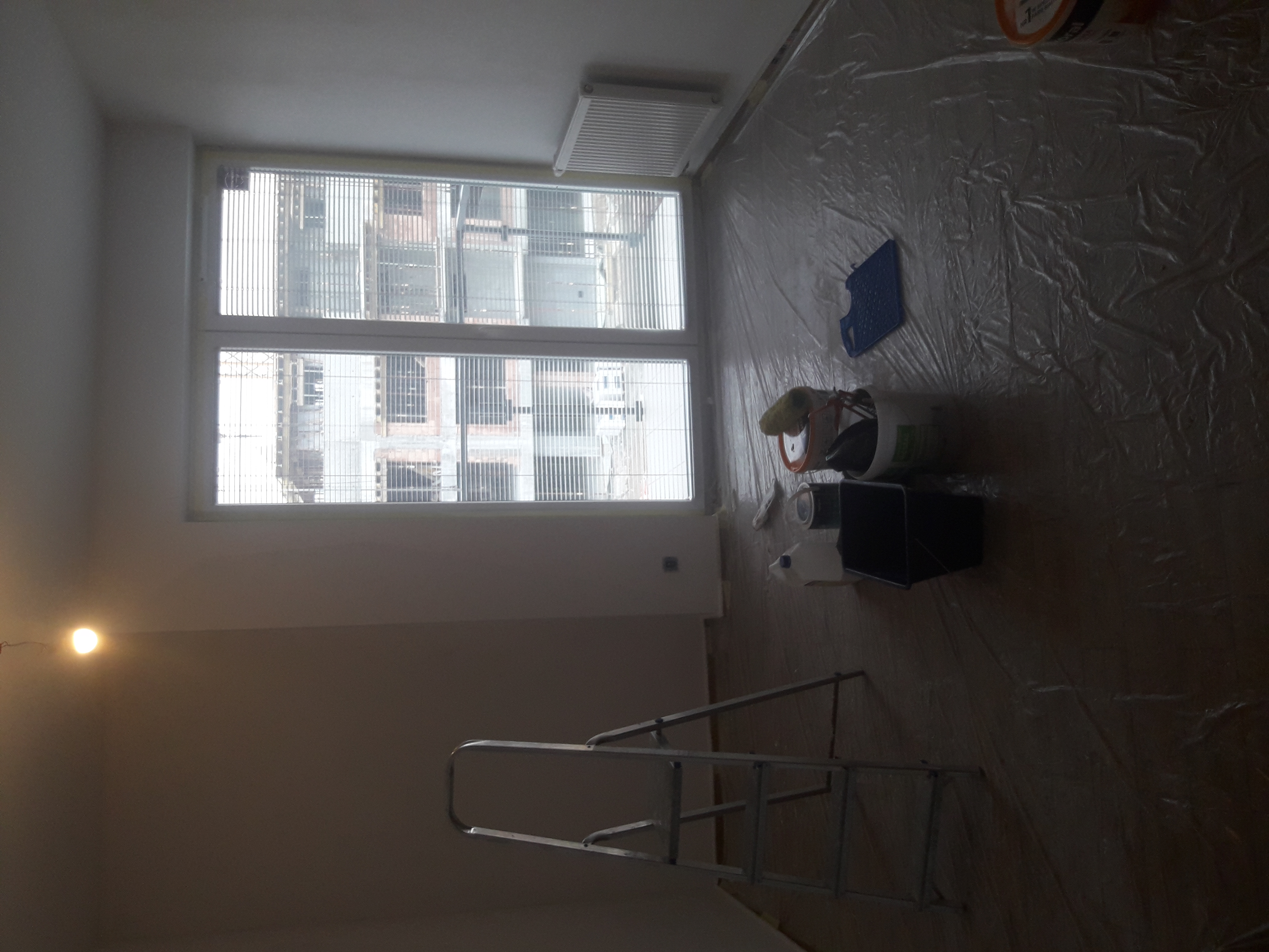 Przygotowania do prac wykończeniowych w pokoju: folia ochronna na podłodze, drabina, wiadro z farbą i akcesoriami malarskimi ustawione przy ścianie z dużym oknem.