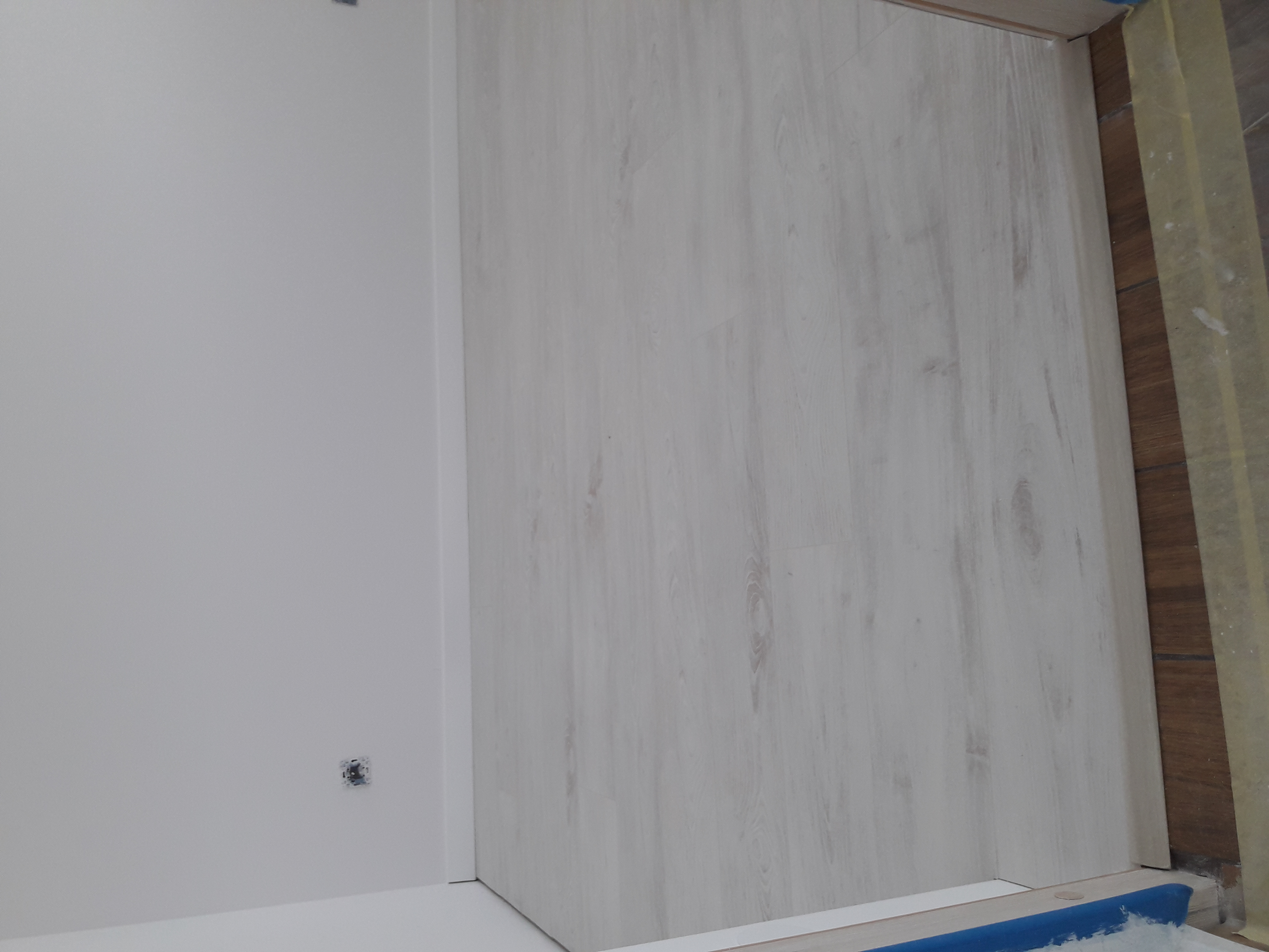 Świeżo pomalowana, biała ściana w rogu pomieszczenia z widocznym gniazdkiem elektrycznym oraz jasna podłoga imitująca drewno, częściowo zabezpieczona taśmą malarską.