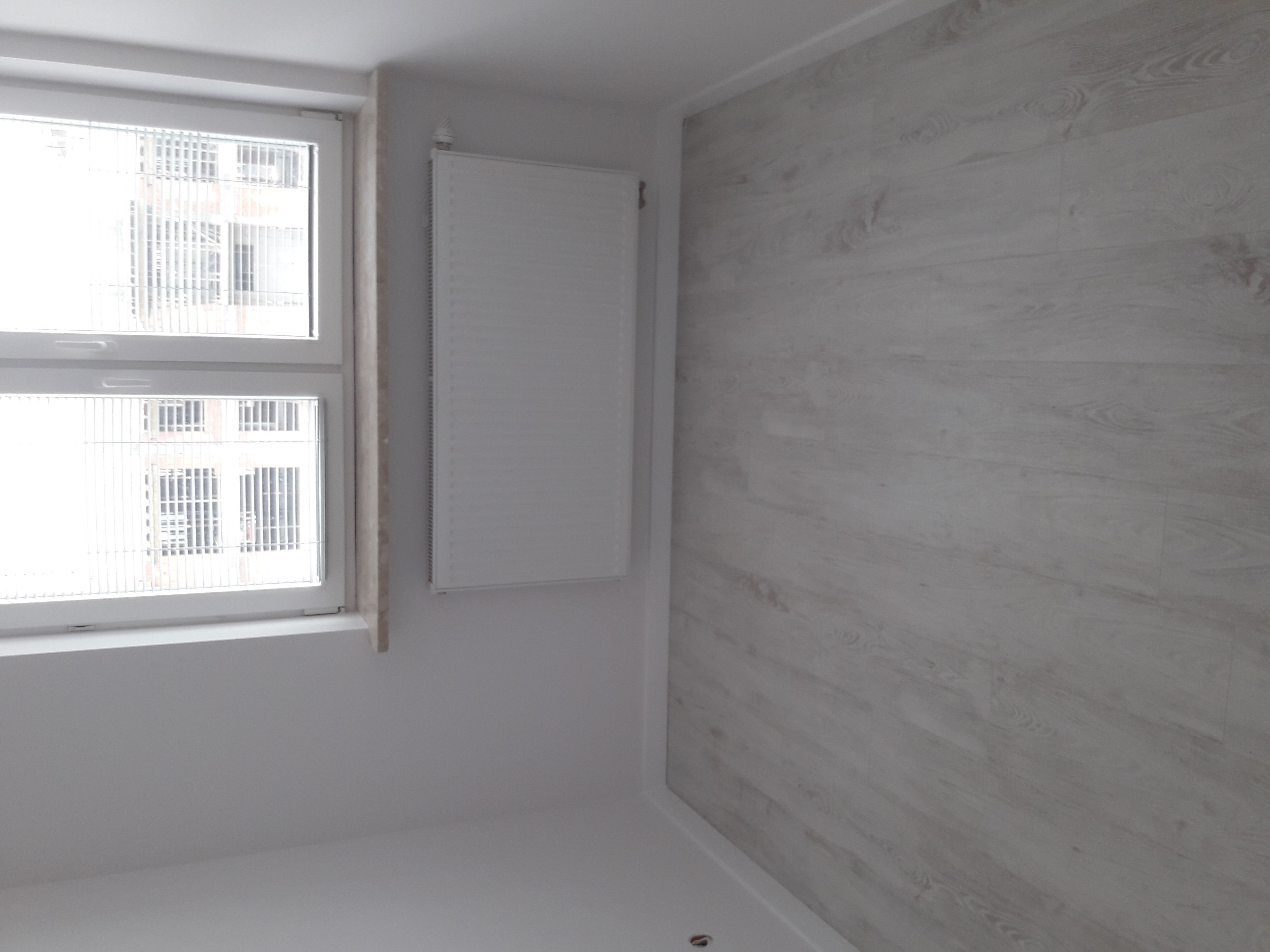 Pusty, jasny pokój po remoncie z panelami ściennymi imitującymi drewno, grzejnikiem i oknem z widokiem na budynek mieszkalny.