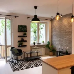 Jasne wnętrze mieszkania z otwartym balkonem, ceglaną ścianą akcentującą, geometrycznym dywanem i wiszącymi lampami w stylu industrialnym.