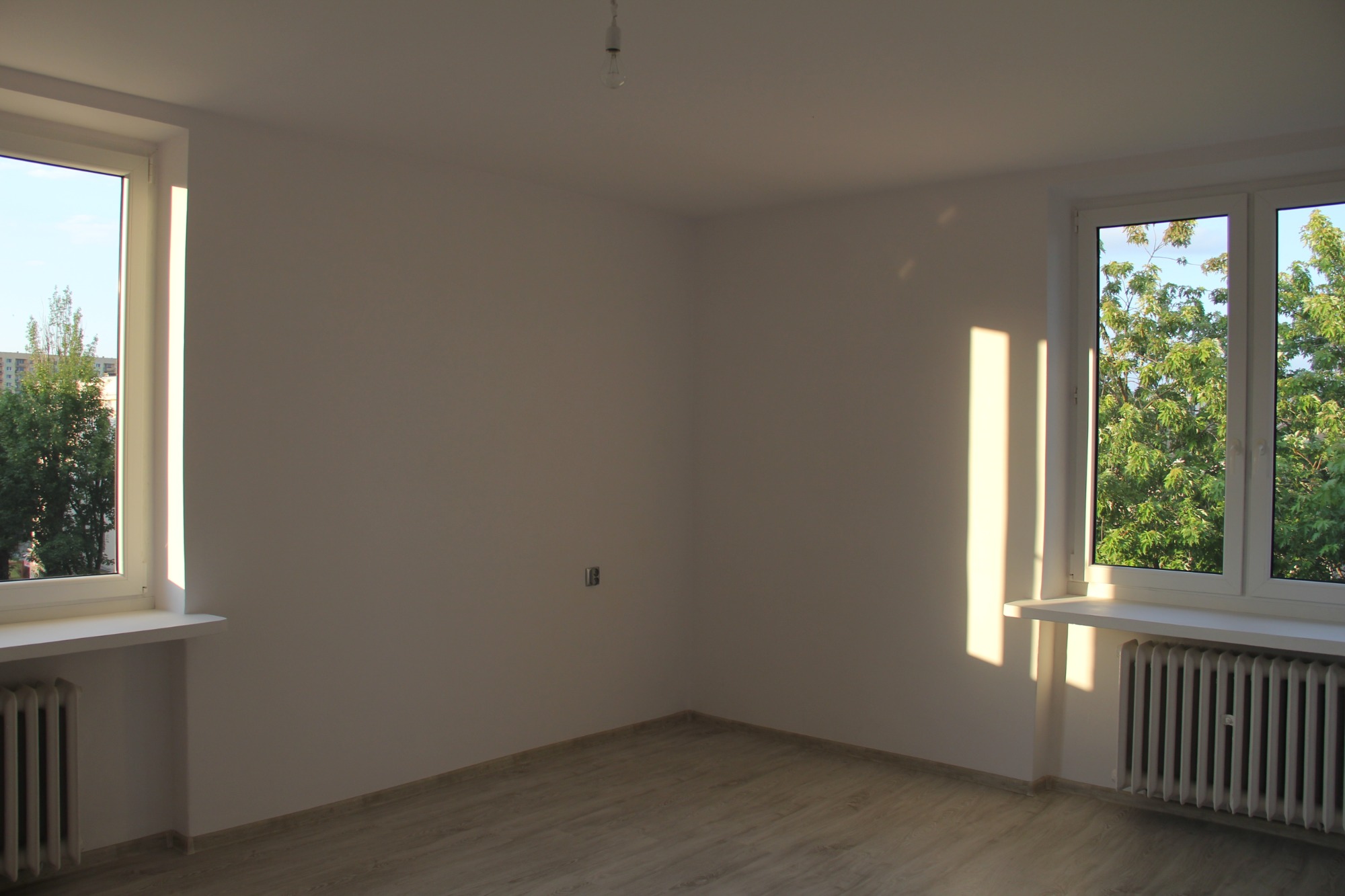Pusty, jasny pokój z dwoma oknami, parkietem i grzejnikami, oświetlony popołudniowym słońcem, z widokiem na zieleń drzew za oknami i wiszącą żarówką bez klosza.