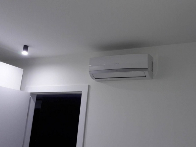 Biały klimatyzator ścienny zamontowany w jasnym pomieszczeniu z widocznym oświetleniem punktowym i fragmentem białych drzwi.