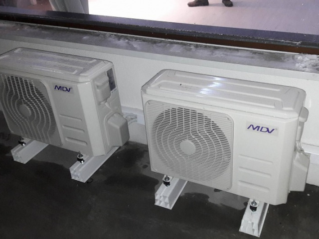Dwa białe klimatyzatory MDV zamontowane na białych wspornikach przy ścianie, widoczne stopy osoby w tle.