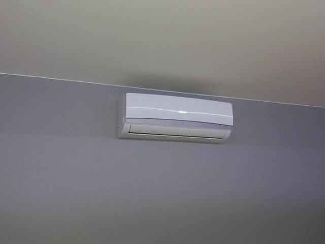 Biała jednostka wewnętrzna systemu chłodzenia powietrza zamontowana na szarej ścianie pod jasnym sufitem, widok z dołu.