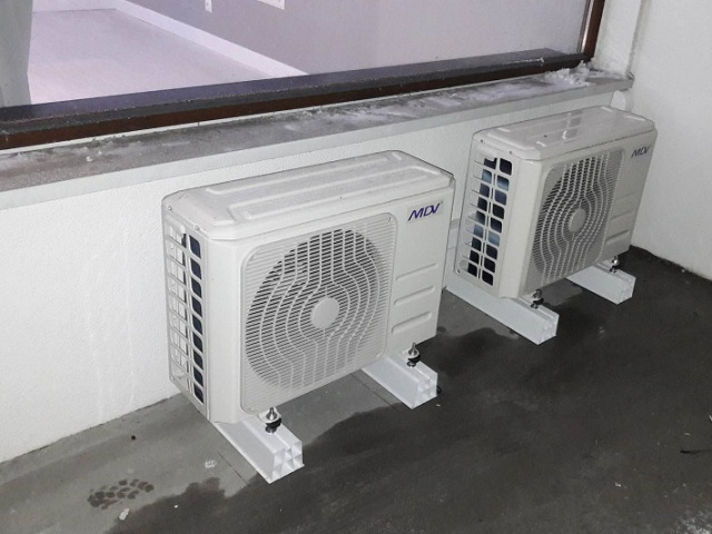 Dwa białe klimatyzatory MDI zamontowane obok okna na białych wspornikach na szarym betonowym podłożu.