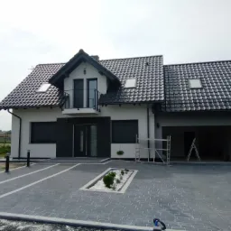 Nowoczesny dom jednorodzinny z szarą kostką brukową na podjeździe, białą elewacją i czarnymi akcentami, w tym balustradą balkonową i żaluzjami okiennymi.