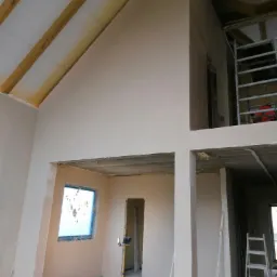 Wnętrze domu w trakcie prac wykończeniowych: świeżo otynkowane ściany w jasnym odcieniu, surowe drewniane belki stropowe, widoczne instalacje, drabina i reflektor budowlany.