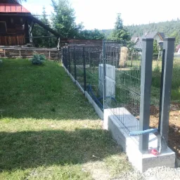 Montaż ogrodzenia panelowego z podmurówką betonową na nierównym terenie, widoczne słupki i siatka, niebieski peszel wystający z podmurówki, w tle domy w stylu góralskim.