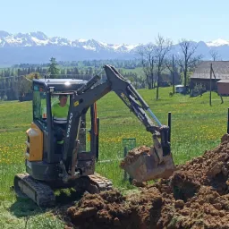 Żółto-czarna minikoparka Volvo z operatorem podczas robót ziemnych na łące z widokiem na Tatry w słoneczny dzień.