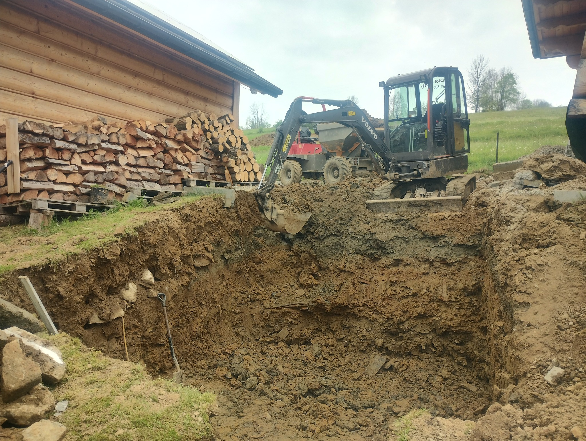 Czarny minikoparka Volvo 633 na gąsienicach, z łyżką zanurzoną w wykopie o pionowych ścianach, obok sterta drewna opałowego przy drewnianym budynku.