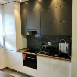 Nowoczesna kuchnia z białymi dolnymi szafkami i ciemnoszarymi górnymi, drewniany blat, czarny zlew i płyta grzewcza, oświetlenie punktowe, widoczny fragment stołu z naczyniami.