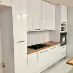Nowoczesna, biała zabudowa kuchenna z drewnianym blatem, czarnym zlewozmywakiem i wbudowanym piekarnikiem oraz mikrofalówką.