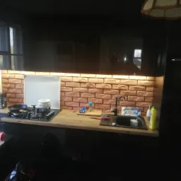 Nowoczesna kuchnia z czarnymi szafkami, blatem z jasnego drewna i ścianą z cegieł, oświetlona listwą LED pod szafkami górnymi.