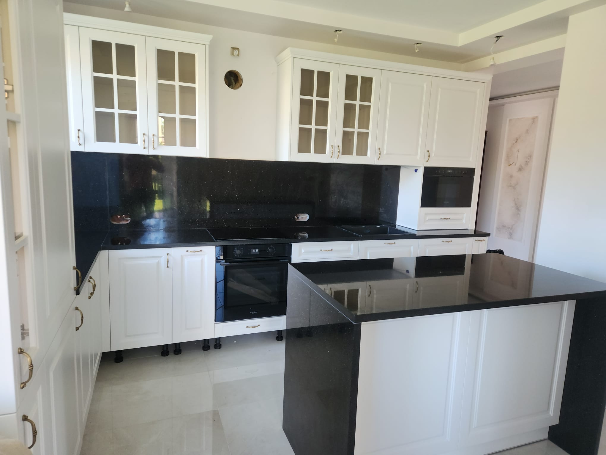 Biała kuchnia z czarnym, błyszczącym blatem kamiennym i wyspą. Widoczne szklane witryny górnych szafek i złote uchwyty. Minimalistyczny, nowoczesny design.