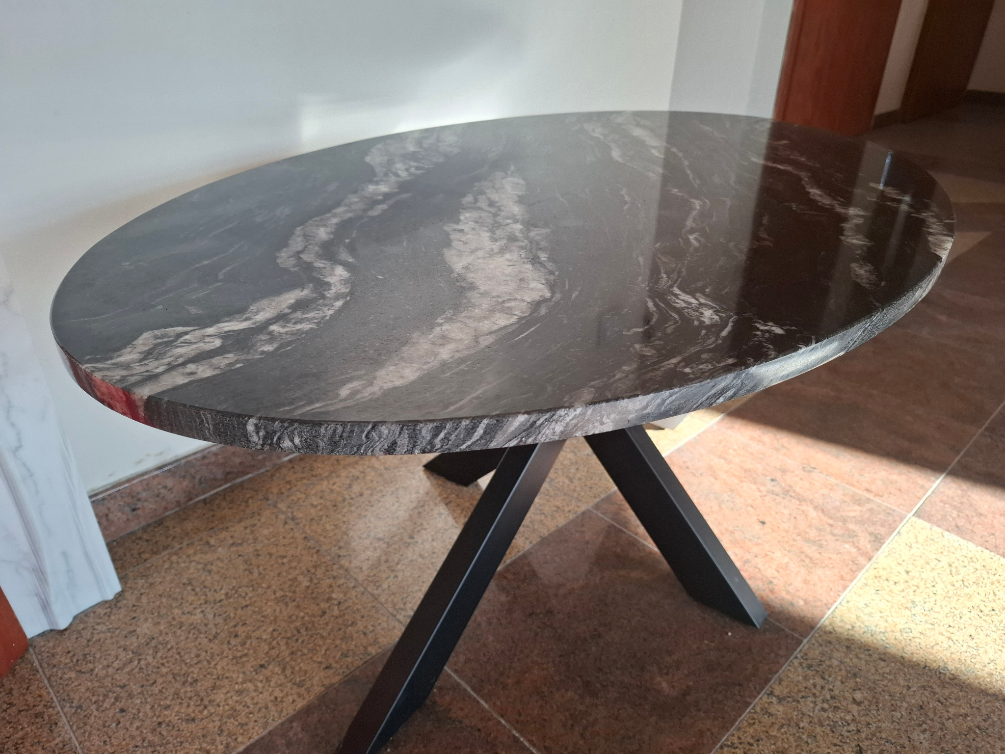 Owalny stolik z blatem z ciemnego kamienia o wyraźnym wzorze, wsparty na czarnej, metalowej podstawie w kształcie litery X. Widoczne detale wykończenia.