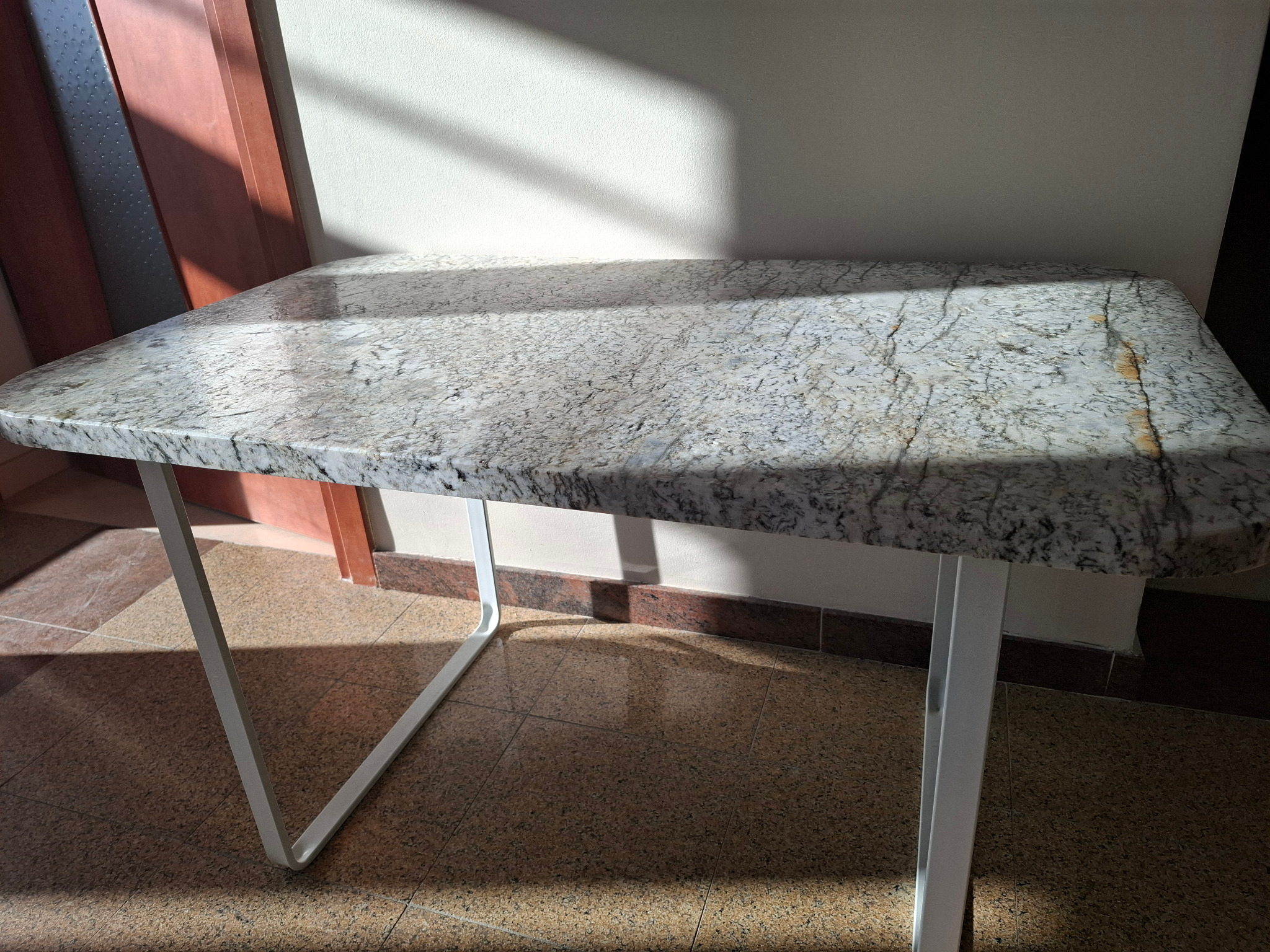 Błyszczący blat stołu z kamienia naturalnego o nieregularnym wzorze, wsparty na białych, metalowych nogach, oświetlony promieniami słońca padającymi na ścianę w tle.