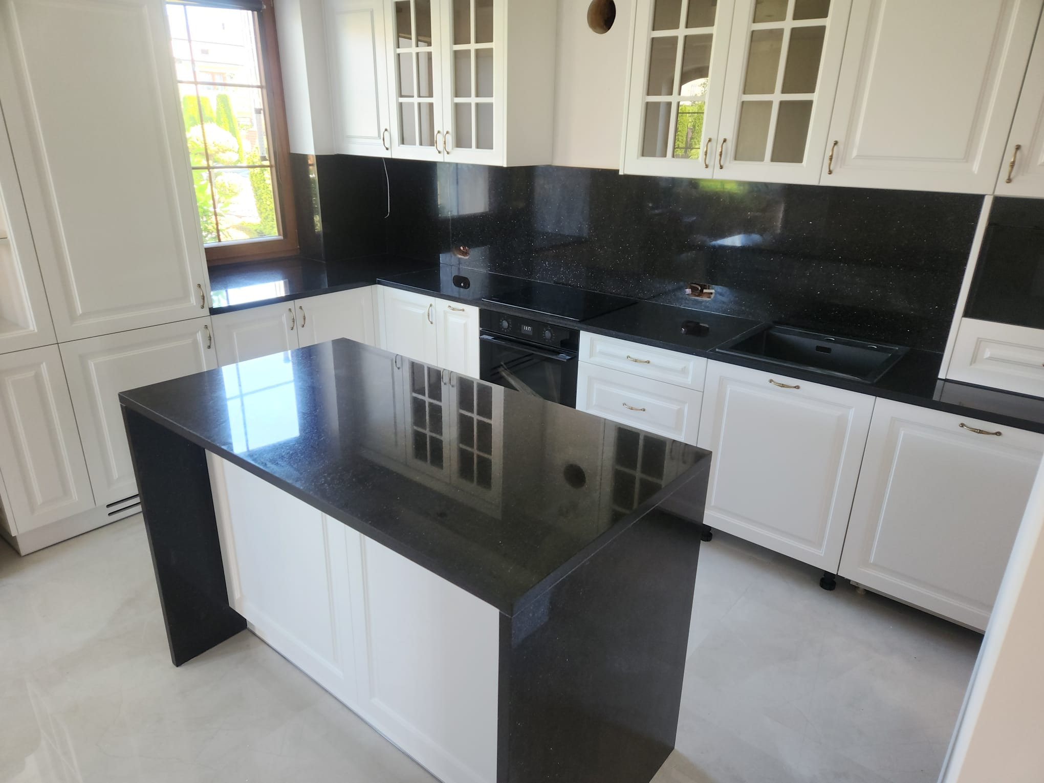 Nowoczesna kuchnia z czarnym blatem kamiennym o błyszczącej powierzchni, białe szafki z przeszklonymi frontami i jasna podłoga. Widok na okno.