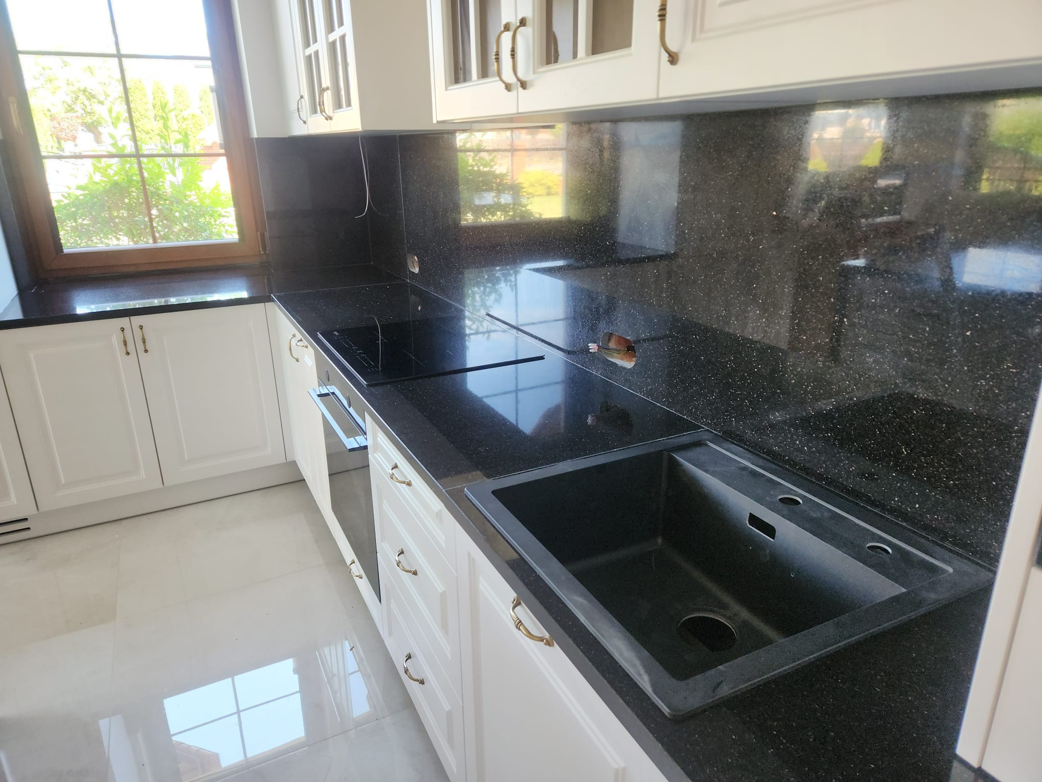 Czarny, błyszczący blat kamienny w kuchni z białymi szafkami i czarnym zlewem. Widoczne okno z drewnianą ramą i fragment płyty indukcyjnej.