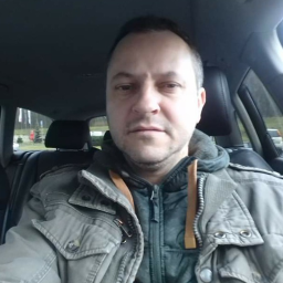 Mwdecor - Autoportret mężczyzny w kurtce wewnątrz samochodu, widok z perspektywy kierowcy, zarys zieleni w tle przez szybę.