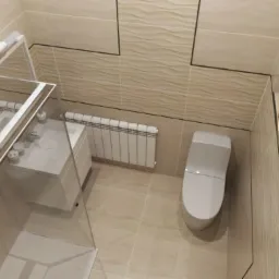 Jasna łazienka z kabiną prysznicową, umywalką z szafką, grzejnikiem i toaletą, wykończona beżowymi płytkami z subtelnym wzorem fal. Wbudowana półka z dekoracyjnymi kulami dodaje charakteru.