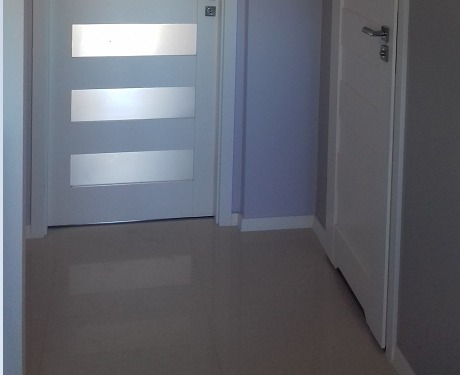 Korytarz z dwiema białymi drzwiami: jedne z trzema poziomymi przeszkleniami, drugie pełne, z prostą klamką. Jasna podłoga z połyskiem, ściany w odcieniach szarości i fioletu.