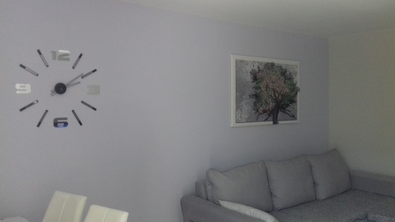 Jasnoszary pokój po odświeżeniu: ściana z zegarem naklejanym i obrazem drzewa nad szarą sofą.