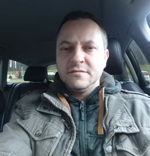 Autoportret mężczyzny w kurtce wewnątrz samochodu, widok z perspektywy kierowcy, zarys zieleni w tle przez szybę.